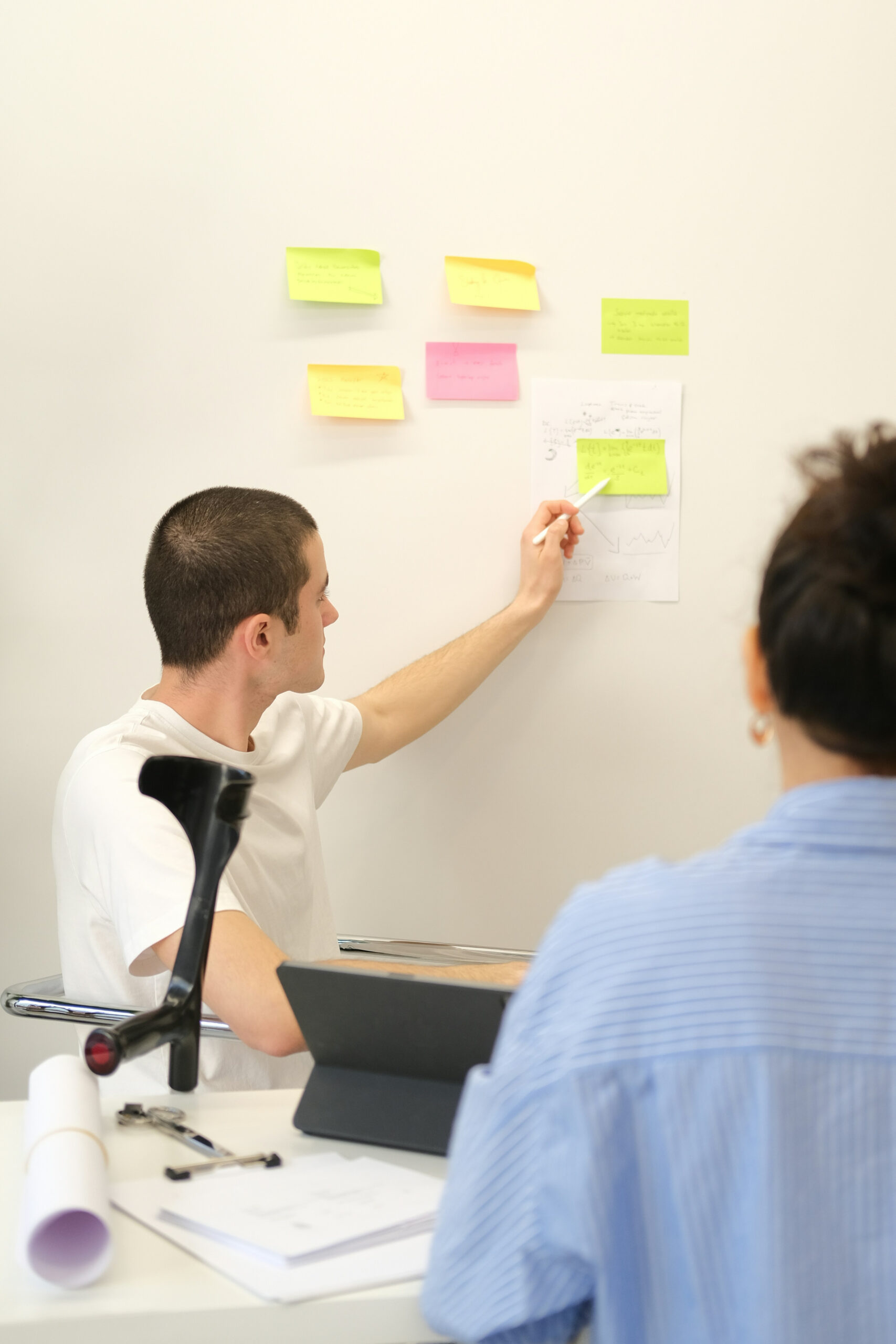 A man in a white shirt from designeo points to a paper and sticky notes on a wall while another person listens in an office setting.