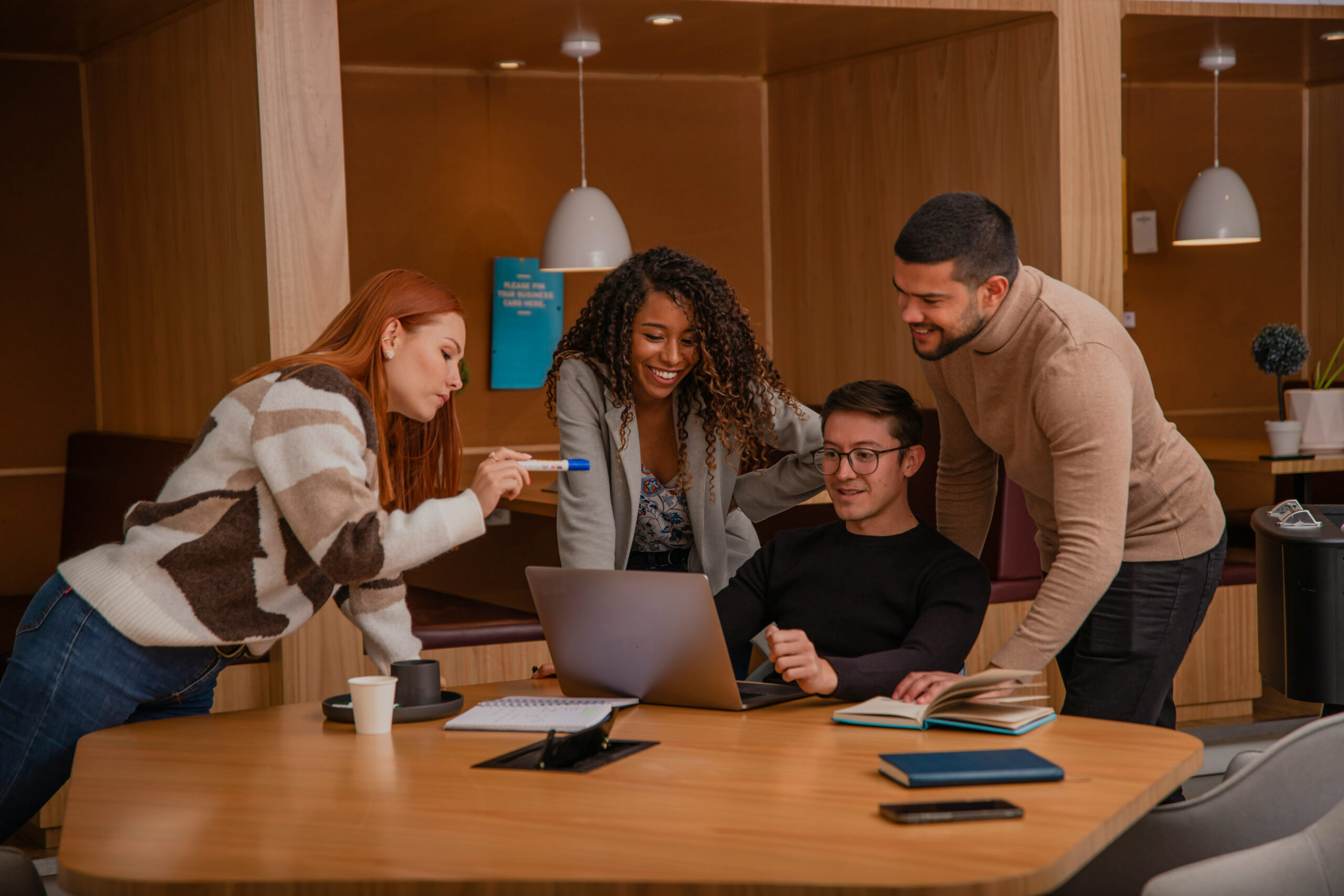 Four people gather around a table in a modern office, smiling and looking at a laptop featuring Designeo, with notebooks, a pen, and a coffee cup on the table.