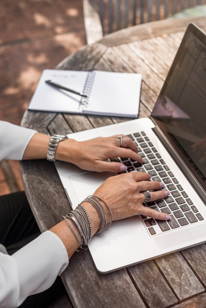 A person typing on a laptop at a wooden outdoor table, with a notebook and pen nearby, working on a designeo project.