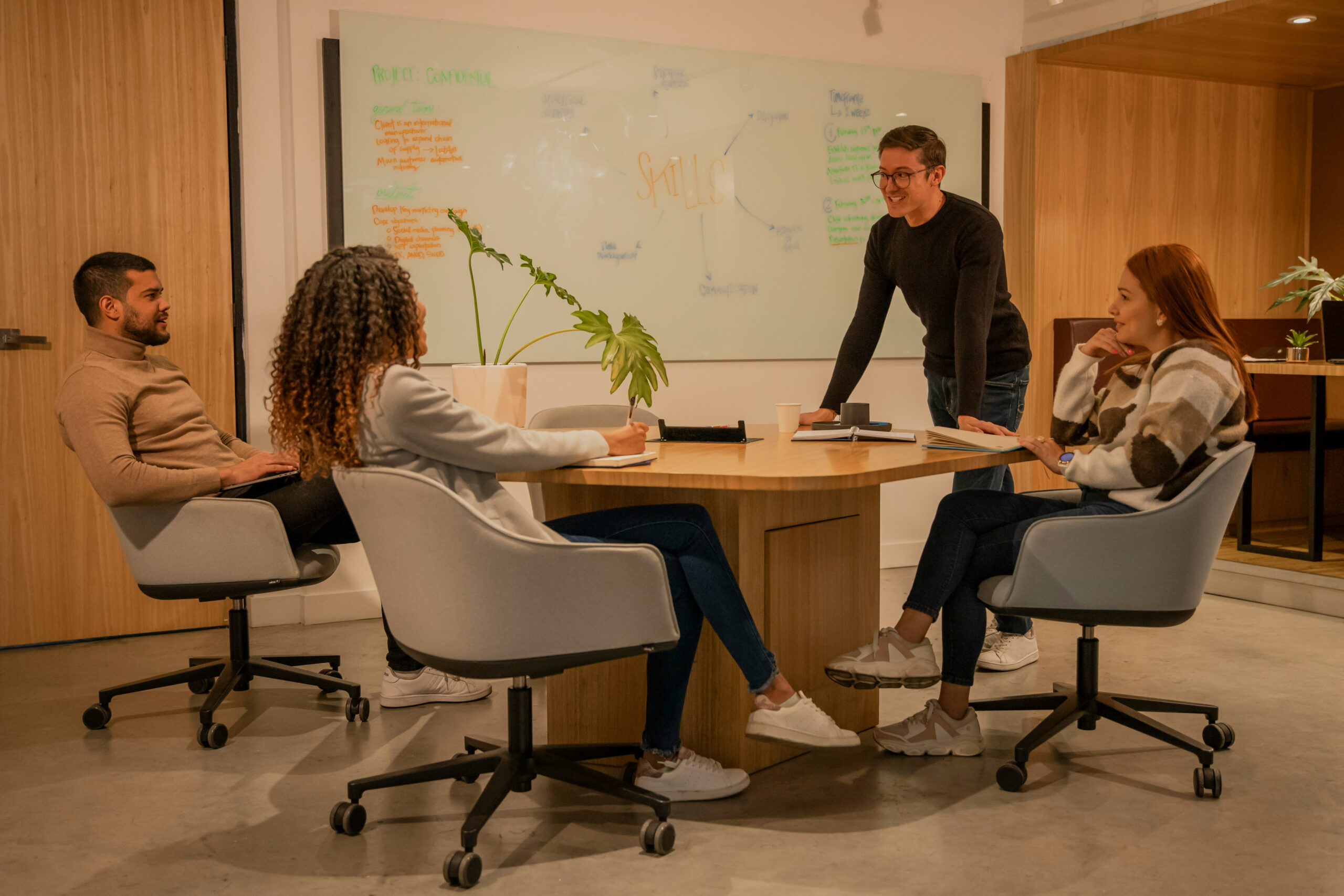 Four people sit around a round table in a designeo office, having a meeting. One person stands and speaks, while the others listen and take notes. A whiteboard with notes is visible in the background.